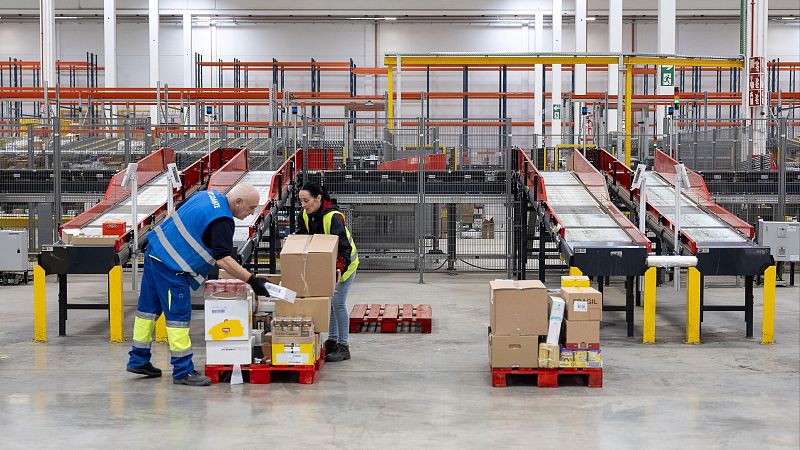Dos empleados, uno con chaleco azul y otro con chaleco amarillo, manipulan cajas en un almacén. Se aprecia un palet rojo lleno de cajas y cintas transportadoras elevadas.