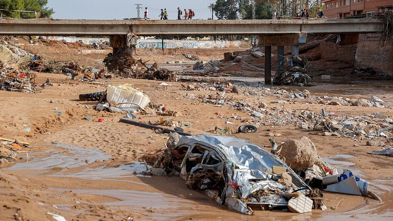 Una rambla desbordada muestra un coche destrozado y escombros, con un puente y personas caminando sobre él. La imagen refleja la devastación causada por una inundación, con edificios y vegetación al fondo.