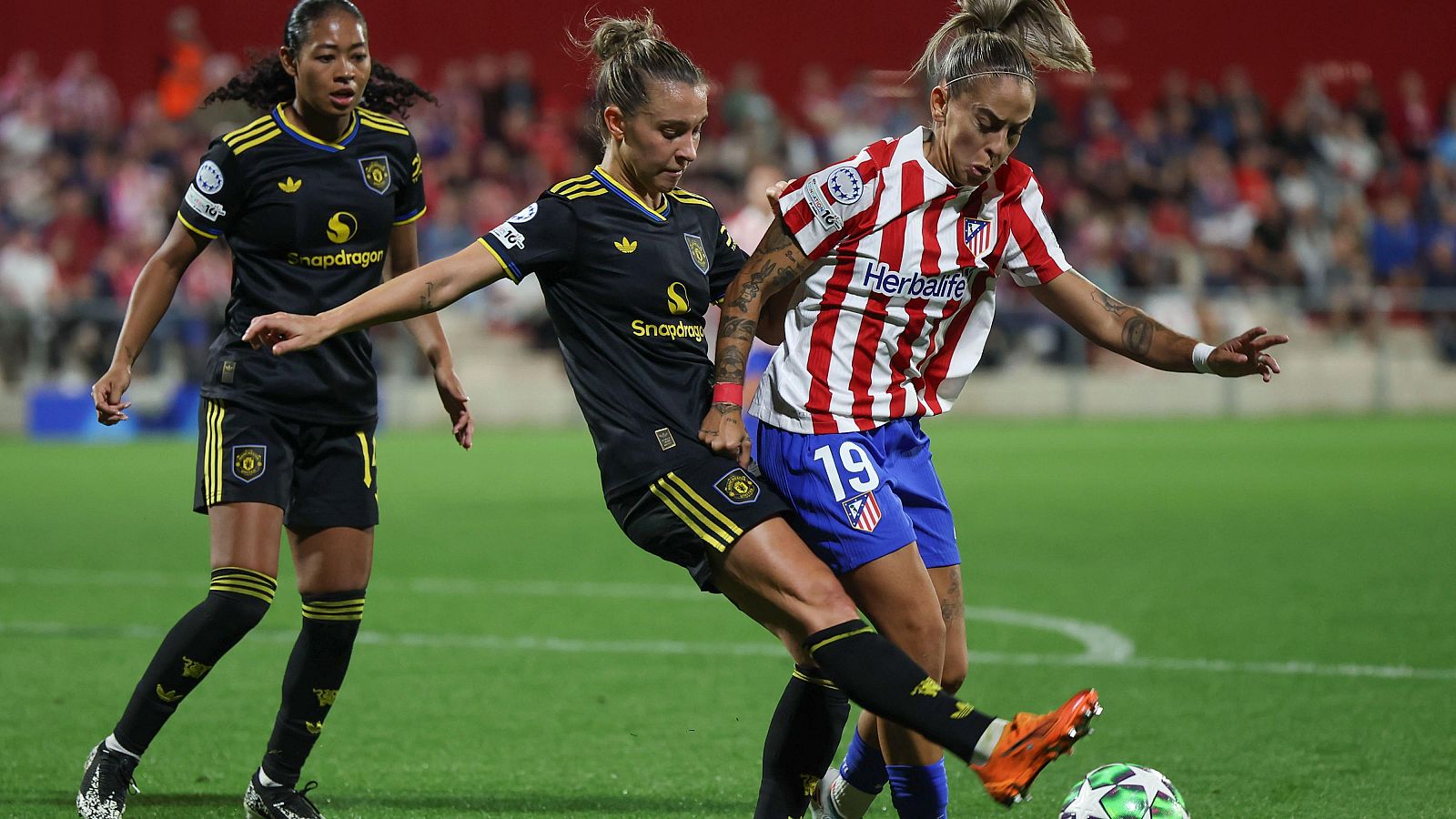 Una jugadora del equipo local, con la camiseta número 19, disputa el balón contra una jugadora del equipo visitante, vestida de negro. El partido se desarrolla en un campo de césped verde, con espectadores en las gradas.