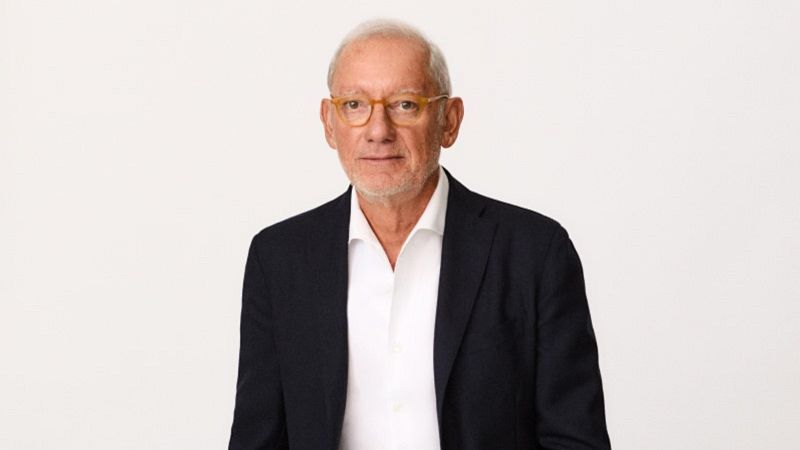 Un hombre de mediana edad, con cabello blanco y gafas, posa con una camisa blanca y una chaqueta azul marino sobre un fondo blanco.