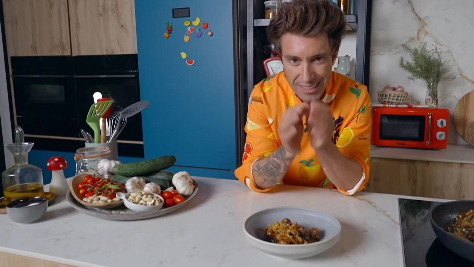 En una cocina, un hombre con camisa estampada y tatuajes presenta un plato de arroz de costilla y bolets, rodeado de ingredientes frescos y utensilios culinarios.