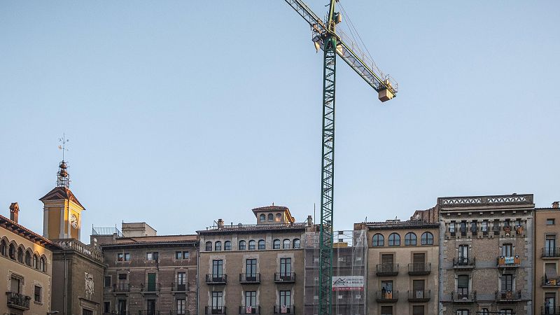 Una vista diurna de Barcelona muestra edificios clásicos con balcones y ventanas, algunos decorados con banderas. Una grúa de construcción se eleva sobre el horizonte, indicando actividad en la zona.
