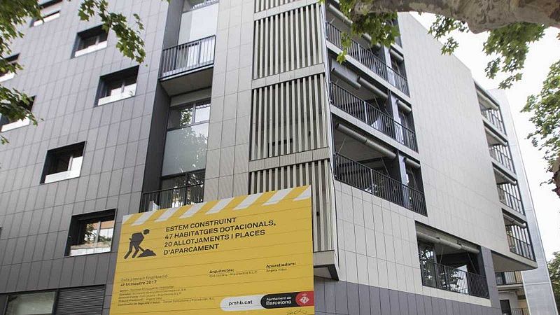Un edificio moderno en construcción con paneles grises y blancos en su fachada, con ventanas cuadradas y balcones. Un cartel amarillo en catalán anuncia la construcción de viviendas y alojamientos.