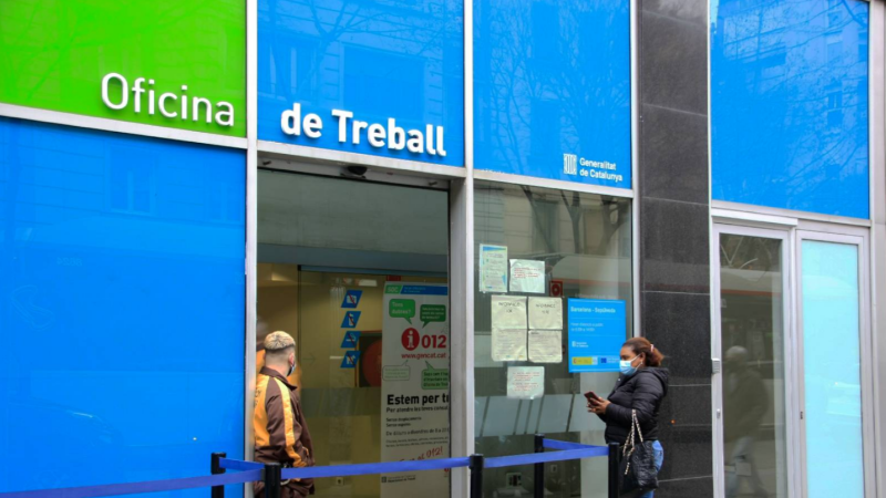 Fachada de una oficina de trabajo con colores azul y verde, con el nombre de la oficina en letras blancas. Hay carteles y avisos en las puertas y ventanas, y una fila con una cinta azul. Dos personas con mascarilla están presentes.