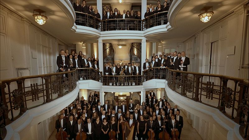 Un grupo numeroso de músicos y cantantes posa en un edificio. Los hombres visten trajes oscuros con pajaritas y las mujeres, vestidos o trajes negros. Se observan instrumentos como violines, violonchelos, arpas y trompetas.