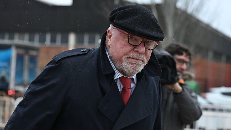 En la fotografía, un hombre con barba y gafas, vestido con gorra y abrigo oscuros, posa al aire libre. Lleva una camisa a rayas y corbata roja, y en el fondo se distingue otra persona.