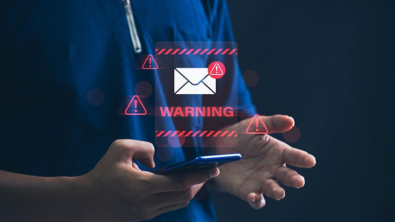 En la pantalla de un teléfono móvil, un icono de correo electrónico con señales de advertencia rojas y la palabra "WARNING" alertan sobre un posible problema de seguridad. La persona que sostiene el teléfono lleva una camiseta azul.