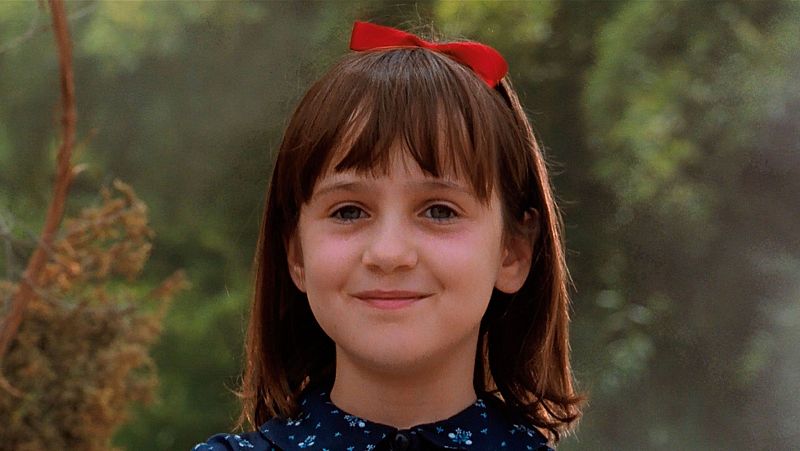 Mara Wilson, como Matilda, posa con un lazo rojo en el pelo y una blusa azul estampada, con una expresión tranquila. El fondo difuminado sugiere un entorno natural.