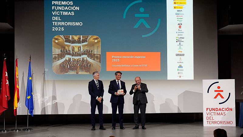 En el escenario de una ceremonia de premios, tres hombres posan frente a una pantalla que anuncia el evento y el reconocimiento a una orquesta y coro. Se observan banderas y logotipos de patrocinadores.
