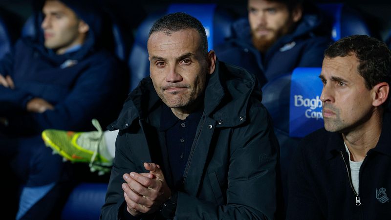 En el banquillo, un hombre con chaqueta oscura y camisa tipo polo oscura está sentado con las manos juntas, acompañado por otra persona con chaqueta oscura. Se distinguen otros miembros del equipo y un logo "Bagh".