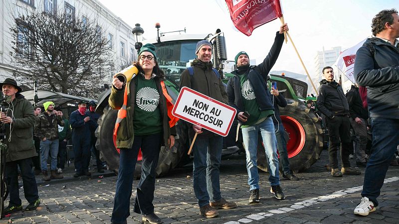Una protesta callejera en Europa muestra a manifestantes con pancartas y banderas. Se observa a una mujer con boina y camiseta verde, y a un hombre sosteniendo una pancarta contra el Mercosur. Varios tractores verdes con ruedas rojas se encuentran en el fondo.