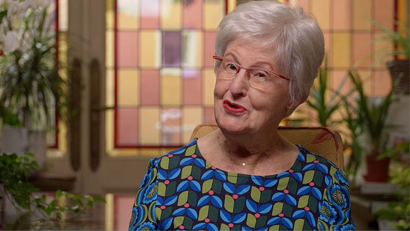 Retrato de una mujer mayor con gafas rojas y labios pintados, vestida con una blusa floreada. El fondo presenta una ventana con vidrieras de colores.