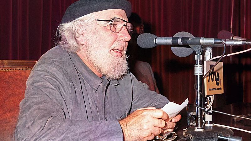 Ernesto Cardenal, con boina y camisa gris, está frente a un micrófono, con un papel en las manos, en un espacio con cortinas rojas.