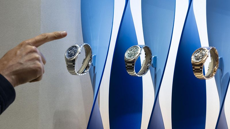 Tres relojes de pulsera, con diferentes acabados en correas y esferas, se exhiben en un escaparate minimalista. Una mano indica uno de los relojes, resaltando su diseño y atrayendo la atención.