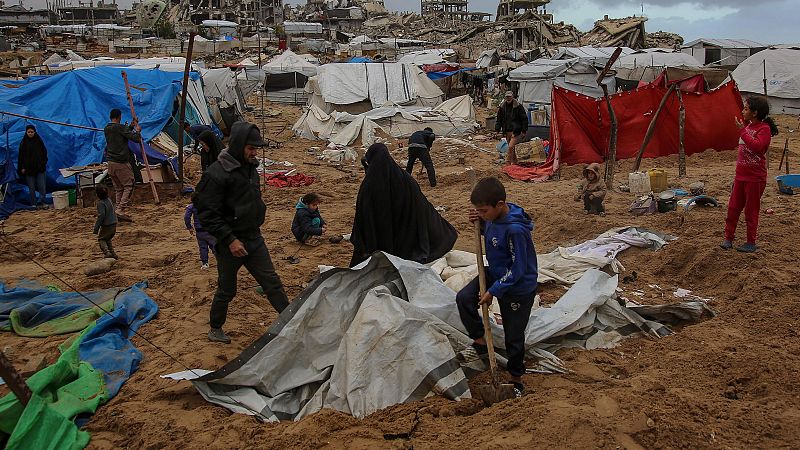 En una zona devastada, un niño con una sudadera azul juega con una pala entre escombros y refugios precarios, mientras una mujer vestida de negro lo observa. El fondo muestra edificios destruidos y más tiendas de campaña improvisadas.