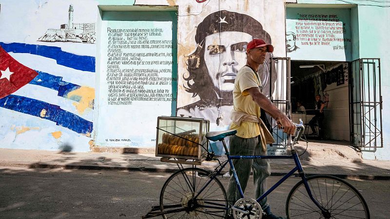 Un ciclista en La Habana, con gorra roja y camiseta amarilla, transporta pasteles en una bicicleta. En el fondo, se observan murales en la pared de un edificio, incluyendo una bandera y un retrato.