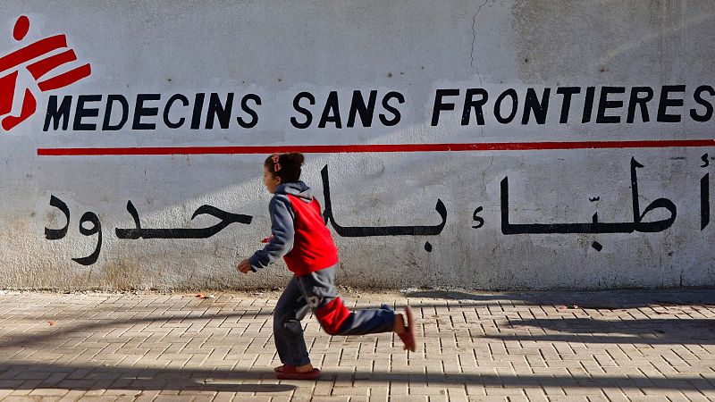 Una niña corre frente a un muro con el logo de una organización humanitaria, vestida con ropa deportiva. El muro muestra el nombre de la organización en francés y árabe, con una línea roja separando los idiomas.