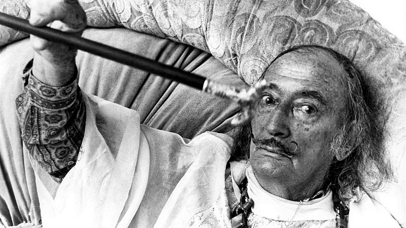Salvador Dalí, con su característico bigote y cabello largo, posa reclinado sobre una almohada. Viste una camisa blanca con encaje y un pañuelo, sosteniendo un puro negro mientras mira fijamente a la cámara.