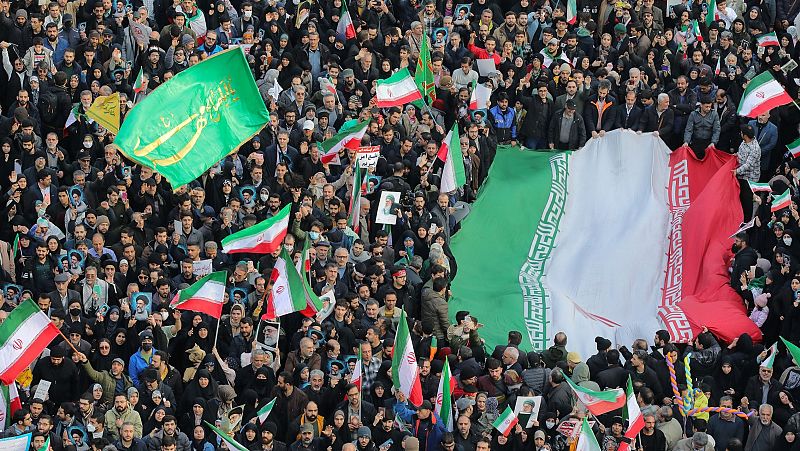 En una plaza de Teherán, una multitud de hombres y mujeres de diversas edades se manifiesta, ondeando banderas y sosteniendo pancartas. La atmósfera general sugiere una manifestación de apoyo.