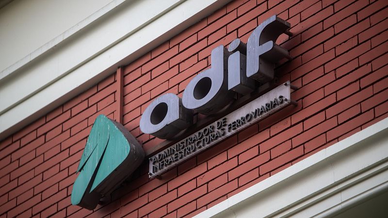 En una pared de ladrillo rojo, se observa un logotipo triangular verde azulado con una forma que recuerda a una "M", junto a la palabra "adif" en letras grises. Debajo, sobre una placa, se lee una inscripción en letras negras.