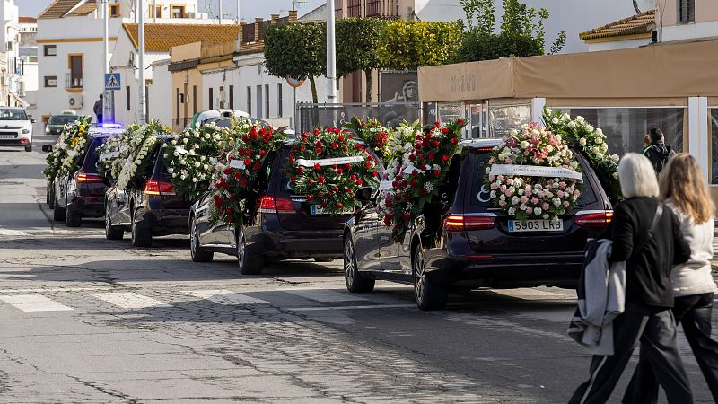 Un cortejo fúnebre con coches adornados con flores recorre una calle urbana. Dos personas caminan en primer plano, mientras que al fondo se aprecian edificios y vegetación.
