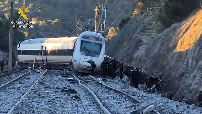 Un tren descarrilado, con la locomotora inclinada y vagones fuera de las vías, es el foco de la imagen. Personal uniformado se encuentra en el lugar, en un entorno montañoso y rocoso, con vegetación escasa.