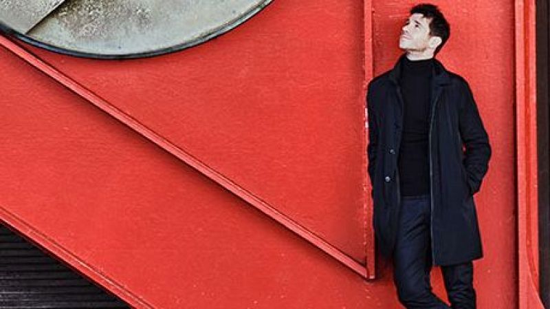 Andrés Salado, con vestimenta oscura y abrigo azul marino, observa algo con atención. El fondo muestra una estructura geométrica roja con líneas que generan un efecto visual.