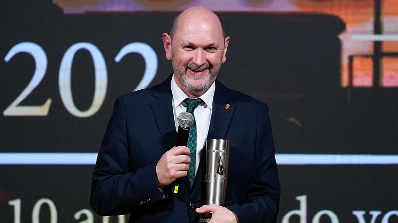 Rafael Louzán, con traje oscuro y corbata, sostiene un micrófono y un trofeo plateado frente a un fondo con números grandes en blanco.