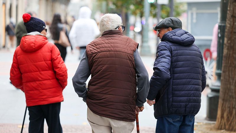 Tres personas mayores caminan por una calle, cada una con un bastón. Uno lleva un gorro azul y chaqueta roja, otro un chaleco marrón y camisa gris, y el tercero una gorra gris y chaqueta azul, todos alejándose de la cámara.
