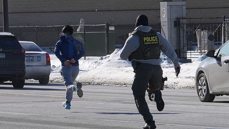 Un hombre esposado huye por una calle cubierta de nieve, perseguido por un agente de policía uniformado. La escena transcurre en un entorno urbano con coches aparcados.