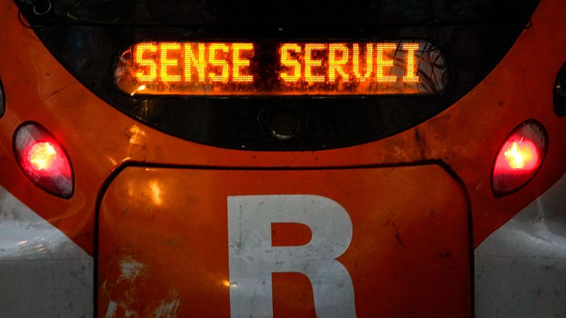 El frontal de un tren naranja de Rodalies exhibe un letrero luminoso con el mensaje "SENSE SERVEI" y el logotipo "R" de la compañía, con luces rojas.