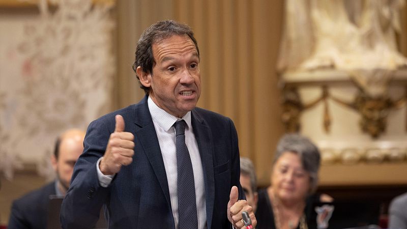 Un hombre con traje azul oscuro, posiblemente un cargo público, gesticula con los pulgares levantados en una sala formal, posiblemente un parlamento o sala de conferencias, con otras personas sentadas al fondo.