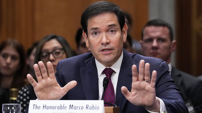 El senador Marco Rubio, con traje y corbata, comparece ante un comité, posiblemente del Senado, con un micrófono frente a él y las manos levantadas, mientras otras personas observan.
