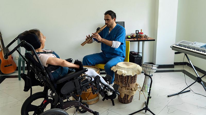 Un musicoterapeuta, en un entorno hospitalario, toca la flauta para una paciente en silla de ruedas. La escena incluye instrumentos musicales como tambores, un teclado y una guitarra, sugiriendo una sesión de terapia musical.