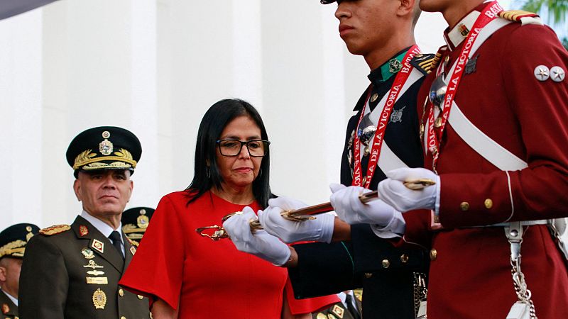 Delcy Rodríguez, con un vestido rojo, está en un acto oficial donde dos militares uniformados sostienen una espada. Otros militares con uniformes similares se encuentran en el fondo de la imagen.