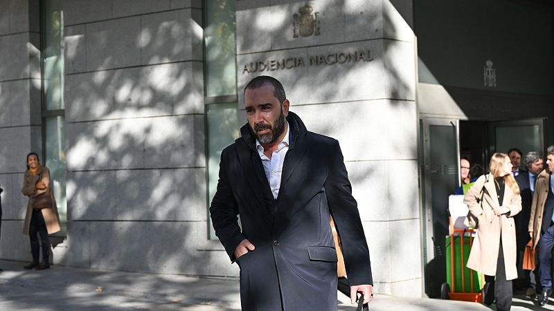 Un hombre con barba y abrigo oscuro, posiblemente De Aldama, sale de un edificio con el nombre "Audiencia Nacional" visible, con una expresión seria y una mano en el bolsillo.