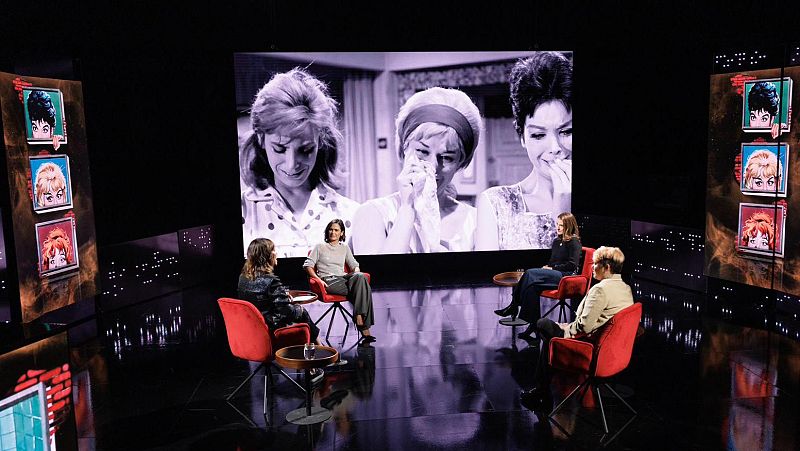 Un estudio de televisión presenta un coloquio con cuatro personas sentadas en sillas rojas, con una mesa auxiliar y un vaso. Al fondo, una pantalla muestra una escena en blanco y negro.