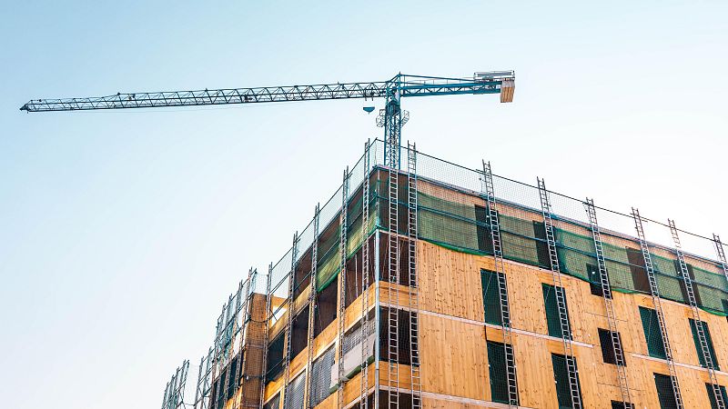 Un edificio en construcción, con estructura de madera clara y ventanas oscuras, se alza contra un cielo azul despejado. La imagen muestra andamios, redes de seguridad y una grúa, indicando actividad constructiva.