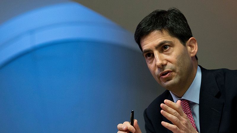 Kevin Warsh, vestido con traje y corbata, gesticula con la mano izquierda mientras sostiene un bolígrafo. El fondo es una pared lisa con una forma ovalada.