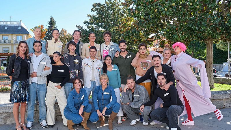 Un grupo de personas posa al aire libre, con etiquetas identificativas. Se observan individuos de diversas edades, con un fondo de árboles y edificios. Una persona llama la atención por su peluca y abrigo rosa.