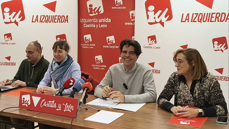 En una mesa, un hombre con jersey gris claro y otros tres individuos participan en un evento. Se observan micrófonos, papeles y un cartel con el nombre de una organización política.