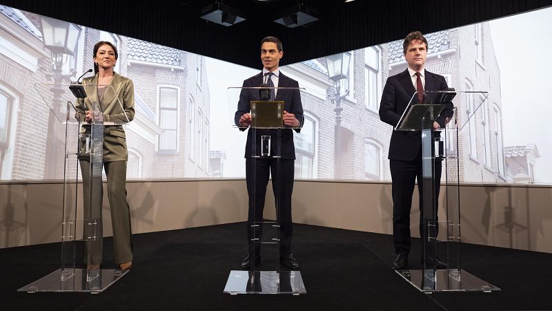 Tres personas, vestidas con trajes, se encuentran detrás de atriles transparentes. Dos de ellos visten trajes oscuros y uno, un traje verde oliva. Al fondo, se proyecta una imagen de edificios.