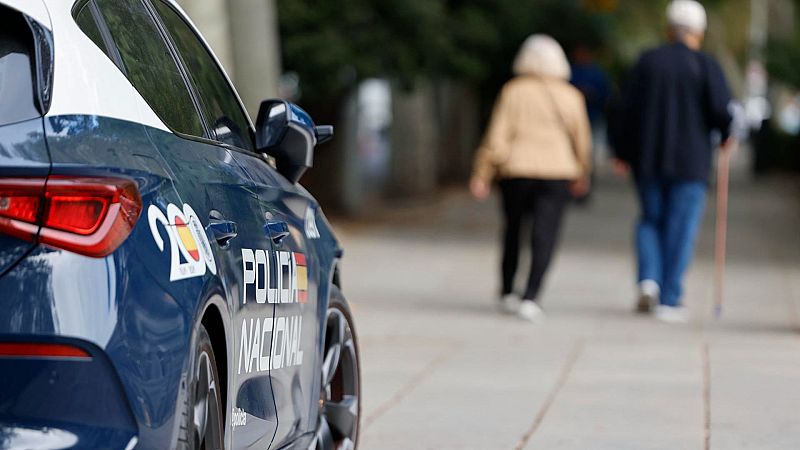 Un coche patrulla de la policía española, con el número 200 en el lateral, se encuentra estacionado. Dos personas mayores caminan por la acera, una de ellas con chaqueta color crema y la otra con chaqueta azul y bastón.