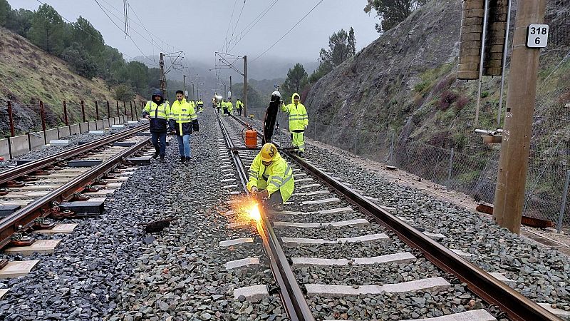 Un grupo de operarios, equipados con ropa reflectante y cascos, trabaja en la reparación de las vías del tren. Uno de ellos está soldando, produciendo chispas, mientras otros supervisan y se mueven por la zona. El entorno es rural y el día está nublado.