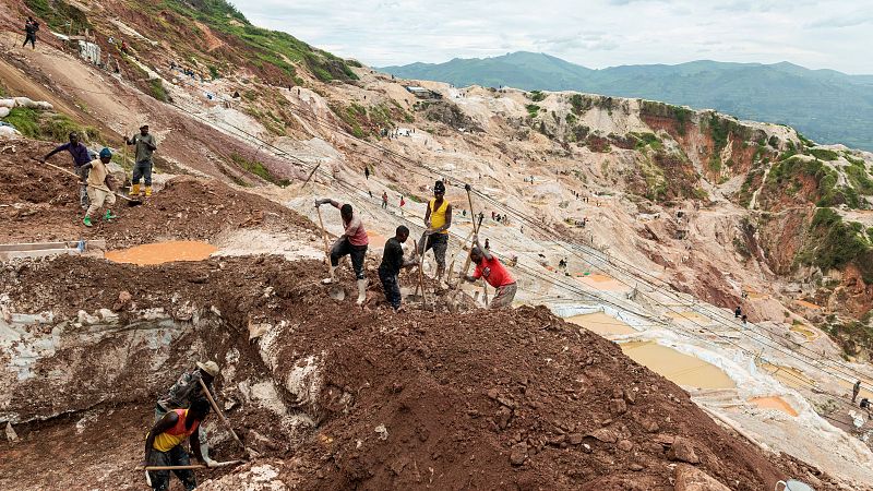 En una ladera montañosa, varios trabajadores excavan con herramientas manuales en un terreno accidentado. El paisaje de fondo muestra montañas con vegetación, sugiriendo una actividad minera en condiciones precarias.