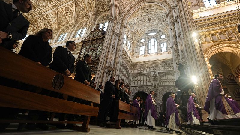 En el interior de una iglesia, se observan personas vestidas formalmente en bancos de madera, mientras una procesión de clérigos avanza por el pasillo, vestidos con túnicas.