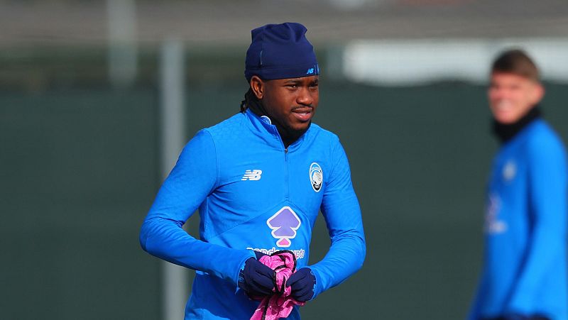 Ademola Lookman, con rastas y gorro azul, sostiene una prenda rosa, vistiendo una chaqueta de entrenamiento azul con el logo de una marca deportiva y el escudo de su equipo. Otra persona con una chaqueta azul similar se vislumbra al fondo.