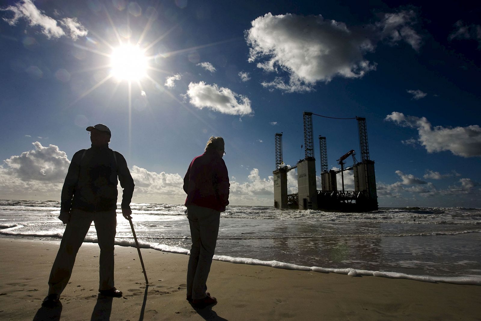 Dos personas observan desde la orilla el dique flotante de la compañía FCC 'Mar del Teide' que ha encallado en la playa de La Barrosa.