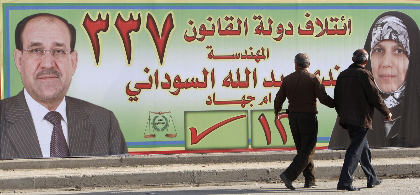 Dos iraquíes pasean junto a un póster electoral que promueve a Nada- al-Sudani y el primer ministro Nuri- al Maliki en Bagdad.