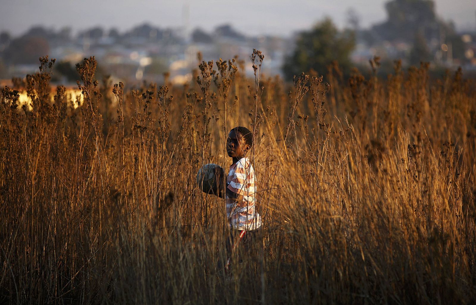 Local child carries a ball while playing soccer at a dirt field in Soweto